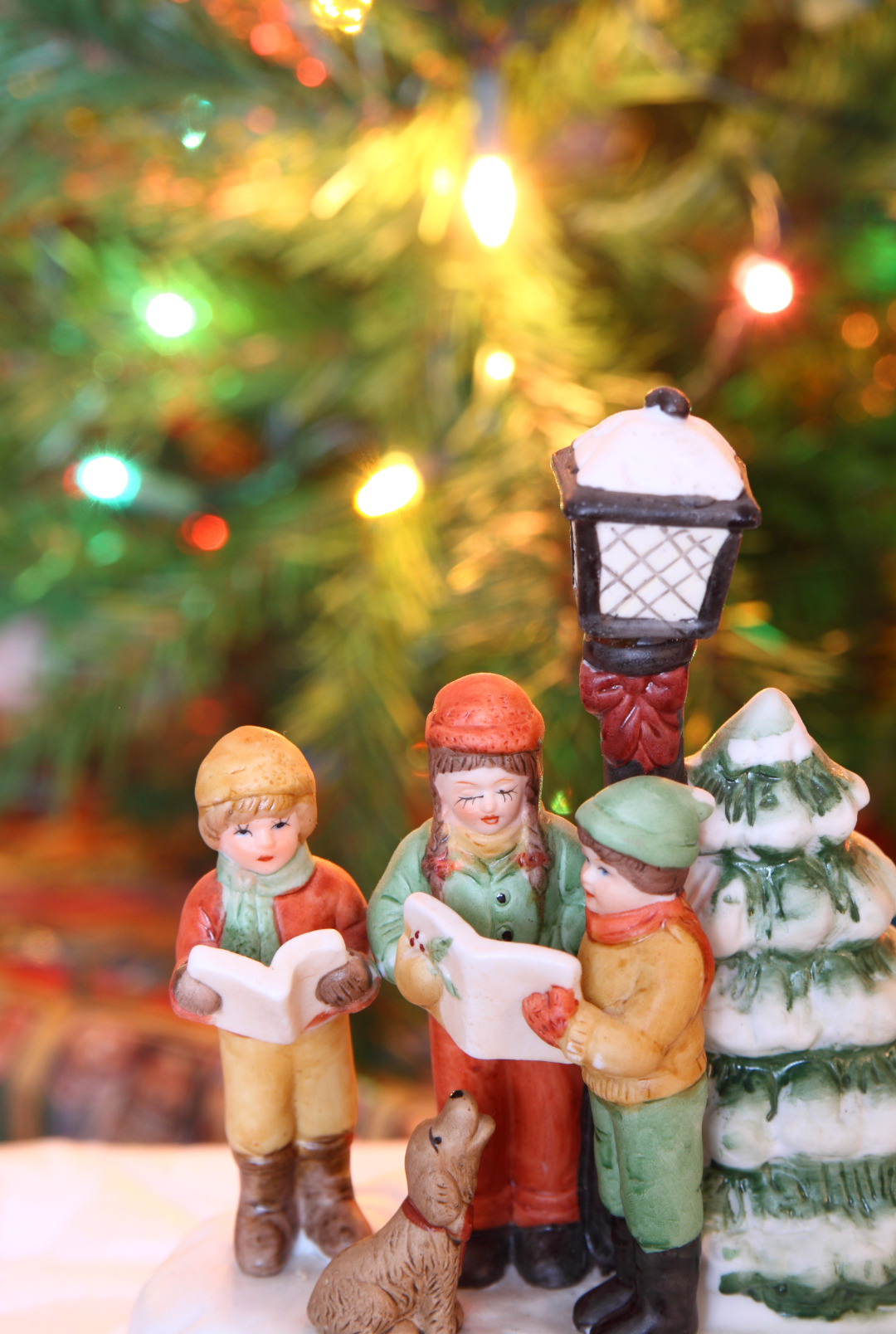 figurines of a girl and 2 boys singing holiday carols. A dog figurine looks up at them. There's a Christmas tree in the background with coloured lights.