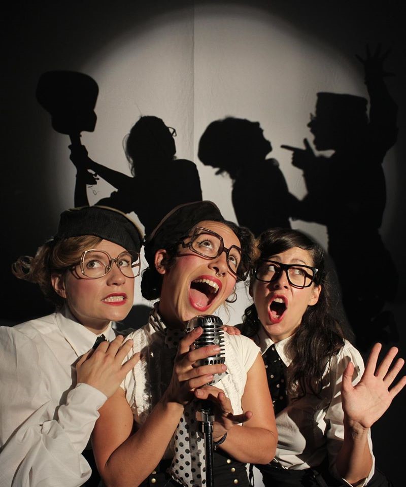 three women surrounding a microphone singing with mouths wide open