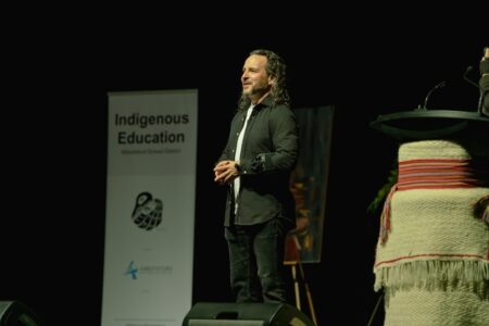 Indigenous storyteller Dallas Yellowfly stands in profile on stage next to a poster that says Indigenous Education.